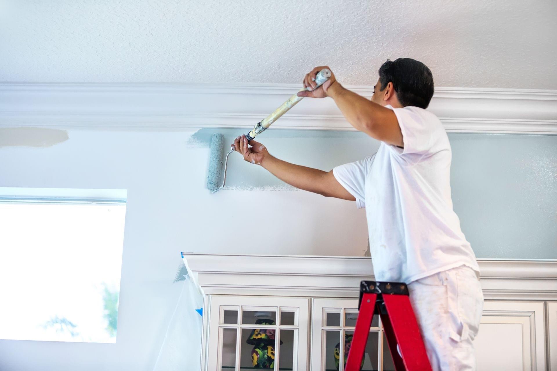 Worker on a ladder rolling light blue interior paint on a wall along crown moulding